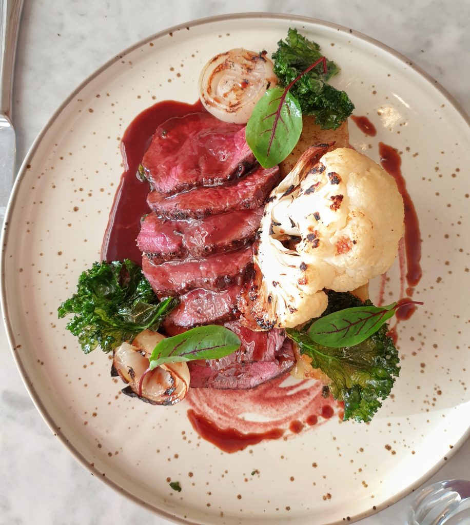 the pan roast loin of venison with baked Roscoff onion, dauphinoise potatoes, chargrilled cauliflower, sautéed rainbow chard, and a chocolate jus 